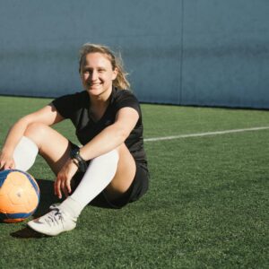 A smiling female soccer player sitting on a soccer field with a ball, enjoying a sunny day.