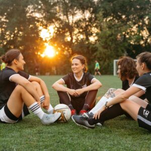 A group of female soccer players sitting on a field, celebrating after a match at sunset.